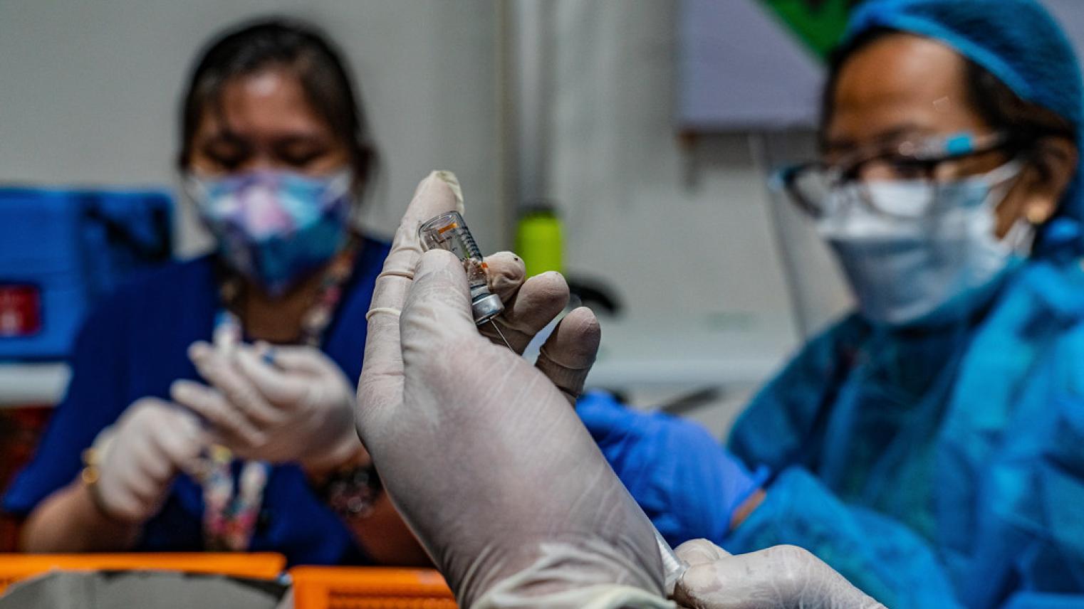 Healthcare workers wear personal protective equipment (PPE) while preparing a vaccine vial and syringe for administration.